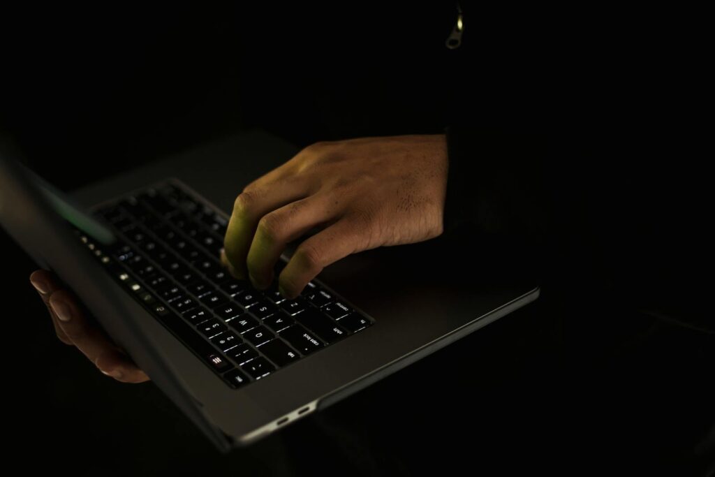 Crop unrecognizable male in black sweater holding netbook on hand and typing on keypad in dark to find videographer in bali
