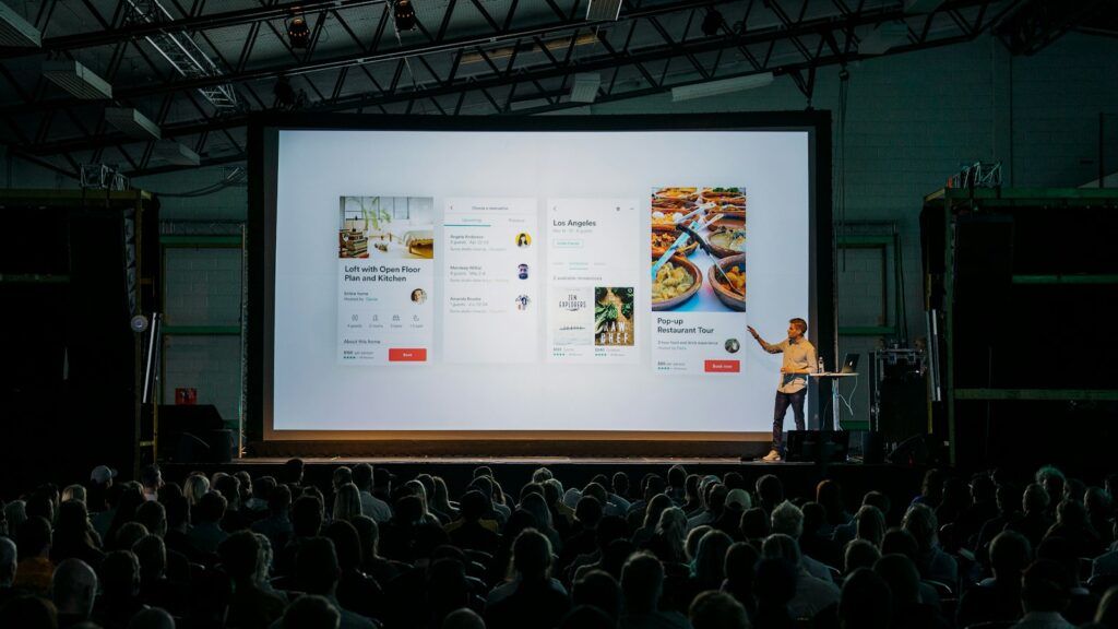 person discussing while standing in front of a large screen in front of people inside dim-lighted room
